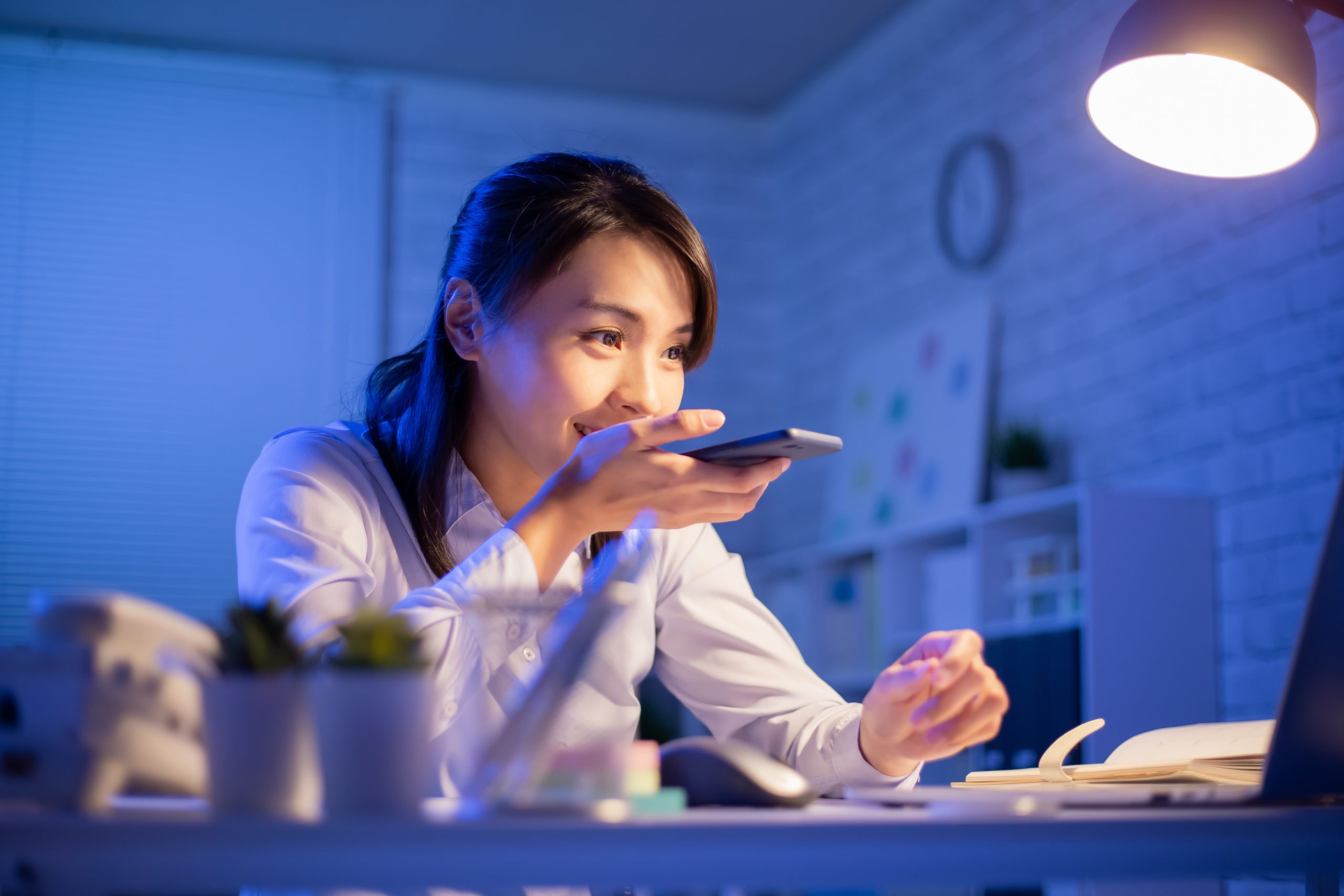 Woman dictating voice notes into her smartphone at a work desk at night.