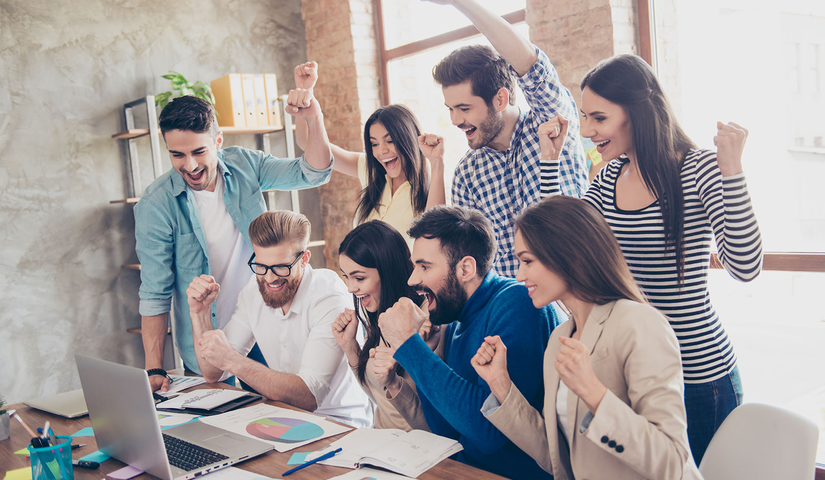 A group of cheerful colleagues in casual clothing, celebrating with arms raised around a laptop in a bright, modern office.