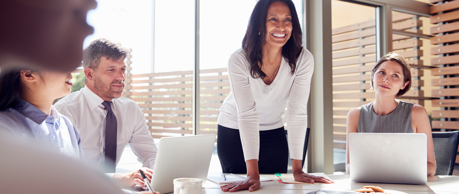 A business meeting in a bright office with a diverse team, a woman standing and leading the discussion.