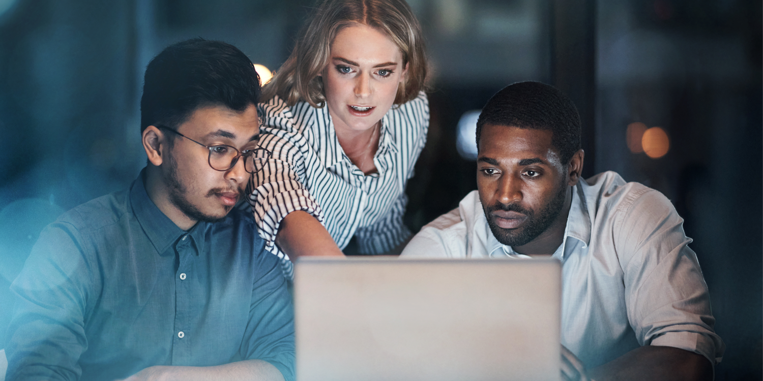 A team meeting at night with three professionals, two men and one woman, focusing intently on a laptop screen.