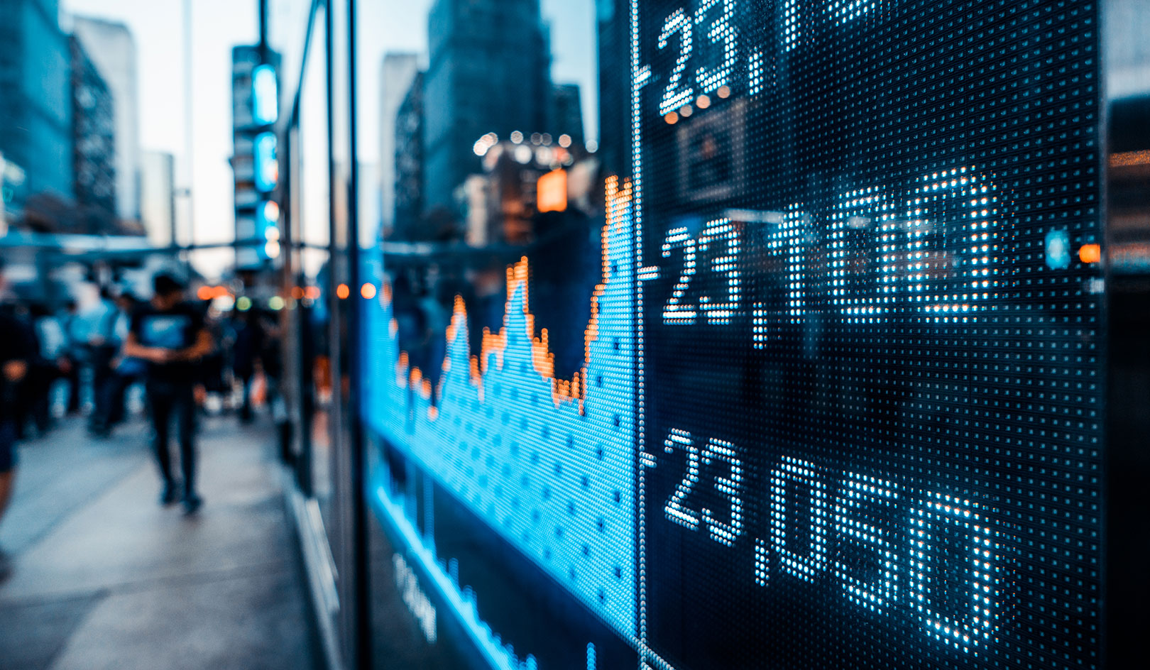 A stock market ticker screen on a city street, showing various stock prices with a blurred crowd of people in the background.