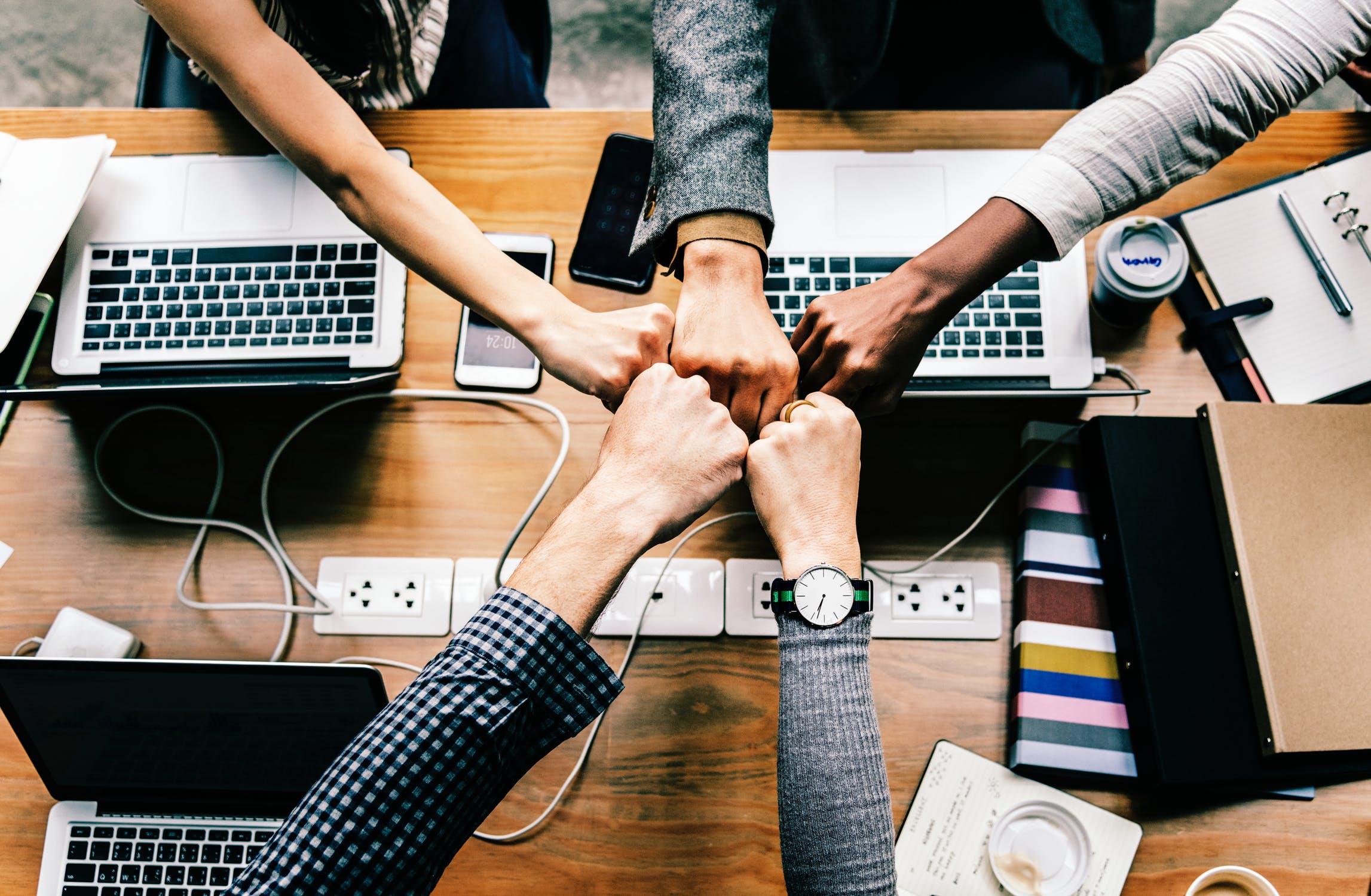 Overhead shot of several individuals in an office setting fist-bumping over a cluttered table with laptops and smartphones.